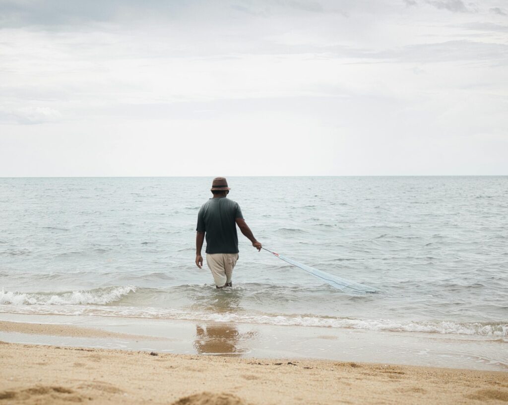 A man in a hat stands in the shallow waves of a sandy beach, holding a fishing net. The water is calm, and the sky is overcast.